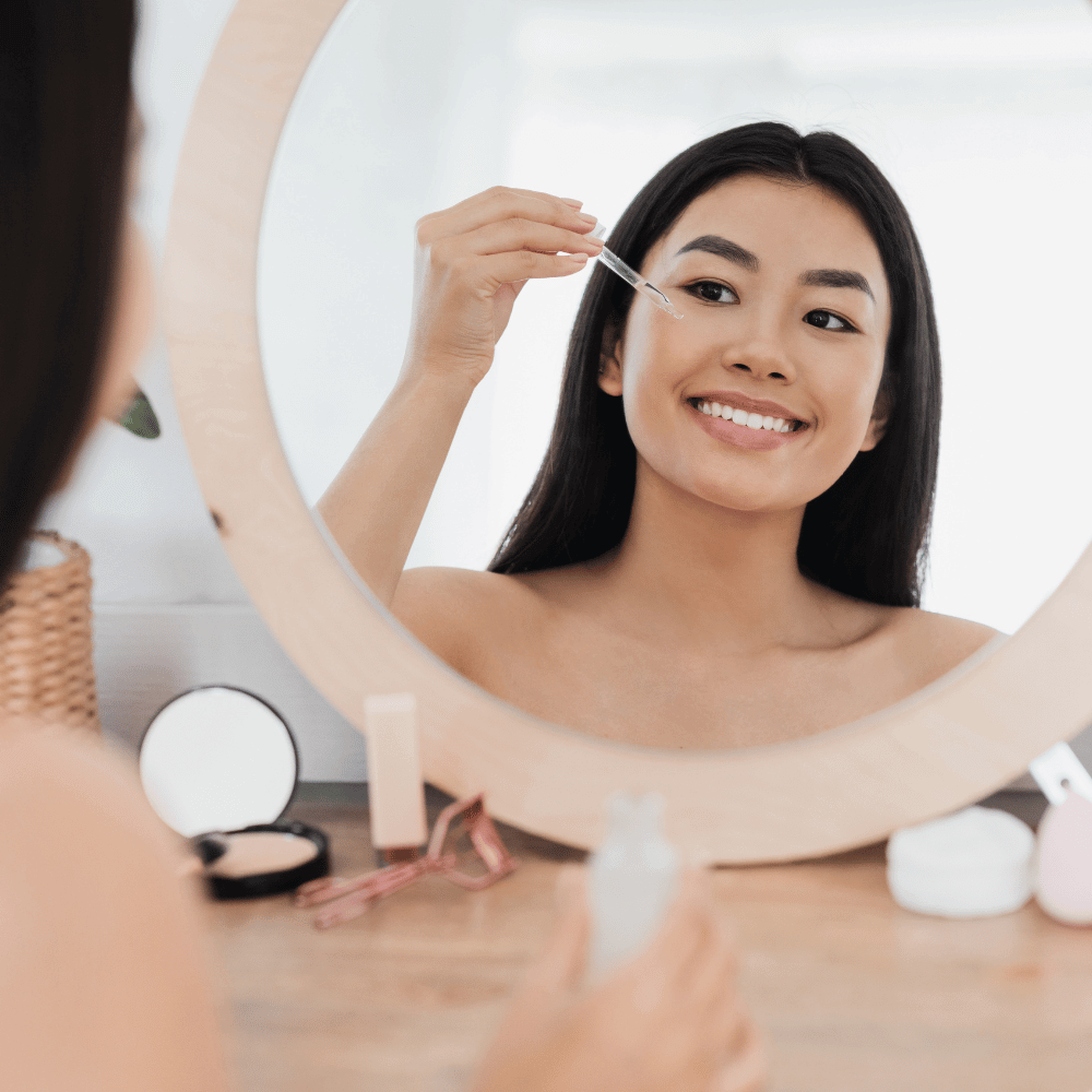 WOMAN PUTTING ON EYE SERUM WHILE SITTING IN FRONT OF HER MIRROR