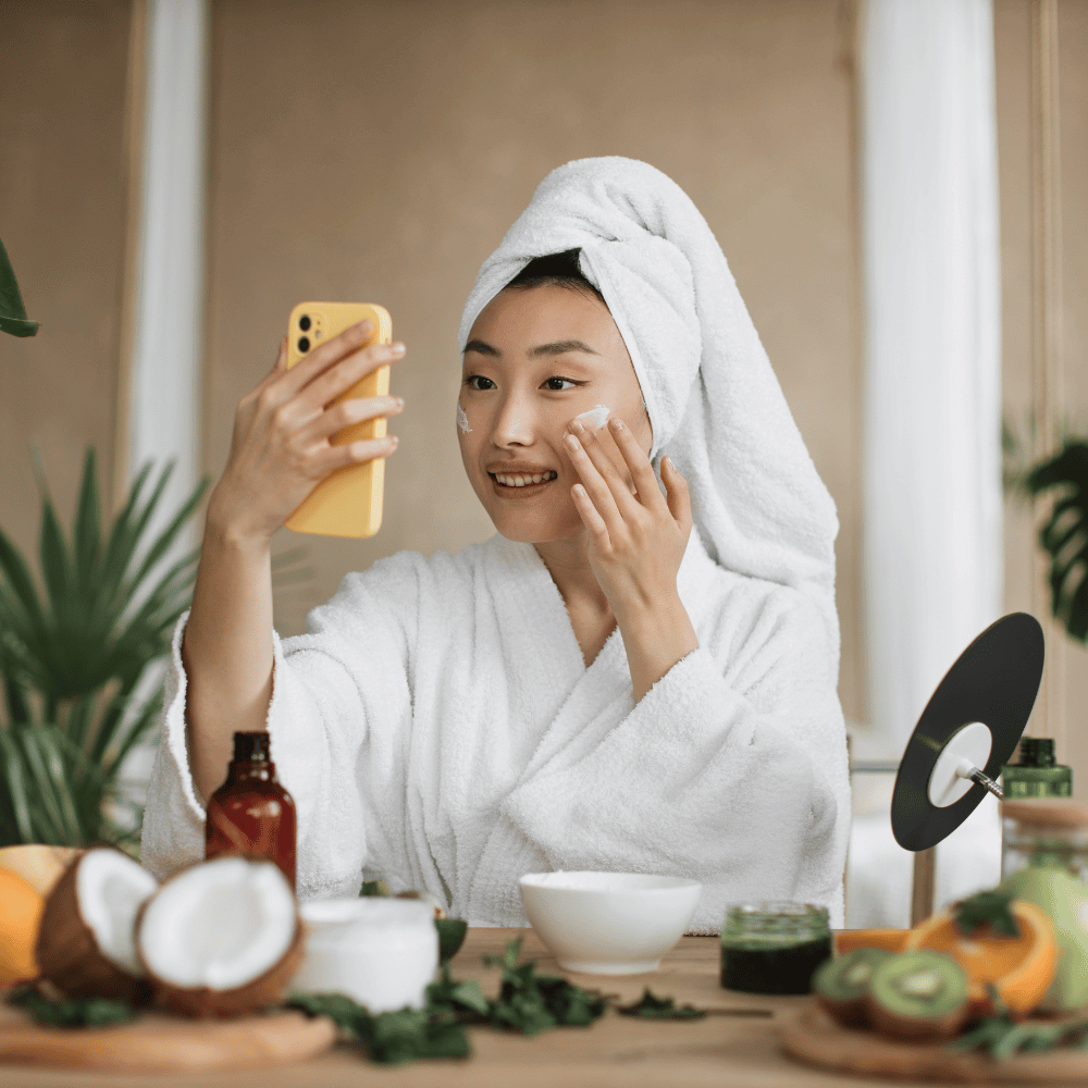 Korean woman in robe surrounded by different natural ingredients and putting on her face